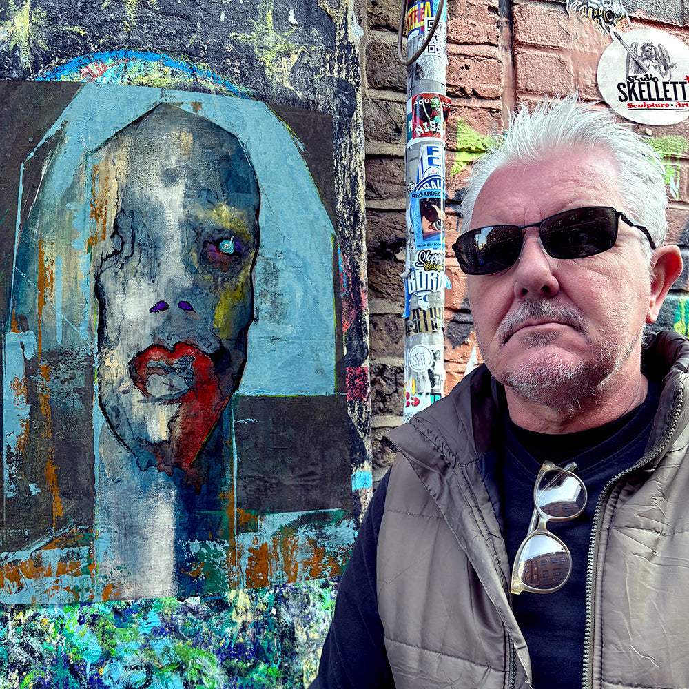 Jay standing next to a graffiti wall with a piece of his artwork and  the Studio Skellett tag.